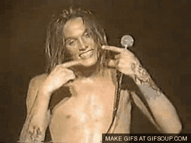 a shirtless man with long hair is smiling and pointing at his face .