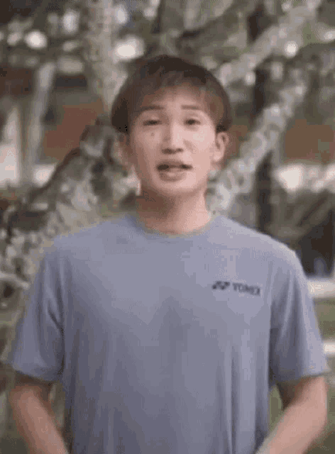a young man in a blue t-shirt is standing in front of a tree and making a funny face .