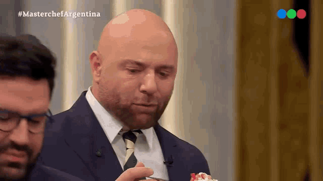 a bald man in a suit and tie is eating a piece of cake with the words masterchef argentina on the bottom