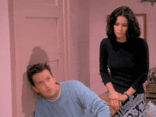 a man in a blue sweater is leaning against a pink wall while a woman stands behind him