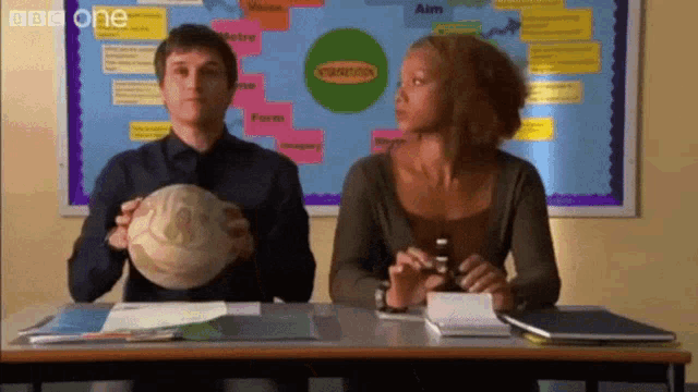 a man and a woman are sitting at a desk in front of a bulletin board that says bbc one