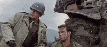two soldiers are standing next to each other in front of a tank in a movie .