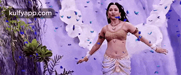 a woman is standing in front of a waterfall with her arms outstretched and flowers falling from the sky .