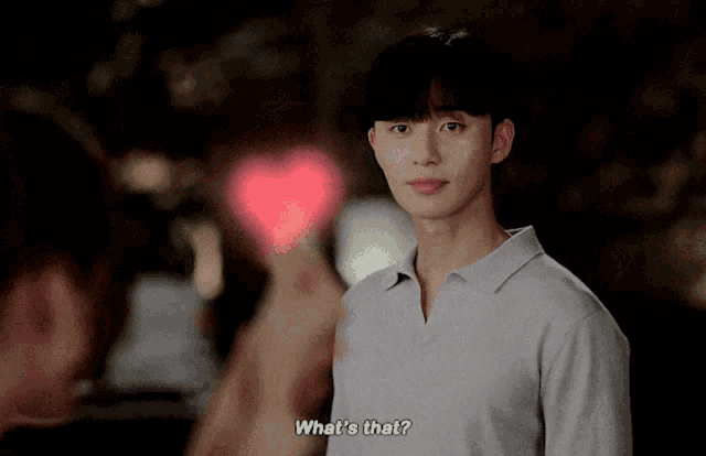 a man in a gray shirt is looking at a woman holding a heart and says " what 's that "