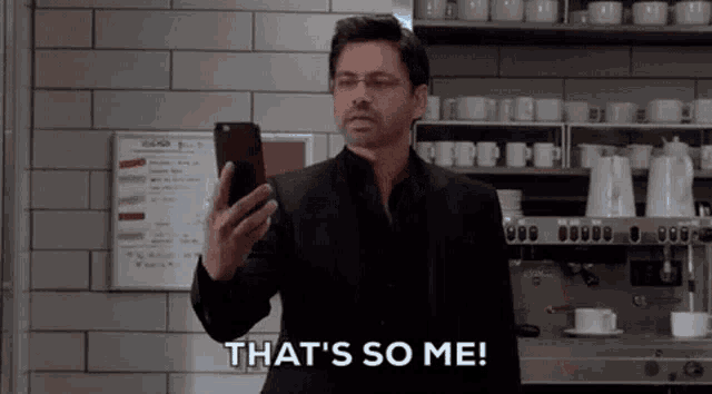 a man in a suit is holding a cell phone in a kitchen and saying `` that 's so me '' .