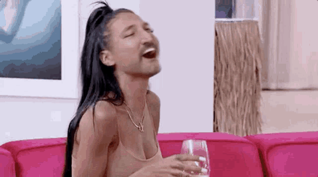 a woman with a mustache is sitting on a pink couch holding a glass of wine .