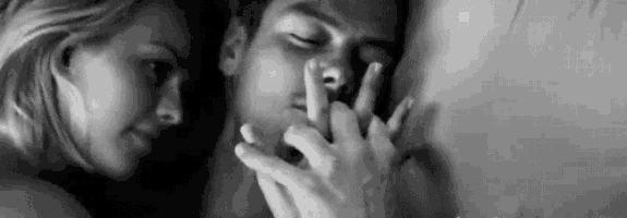 a man and a woman are laying in bed and the woman is touching the man 's face with her hand .