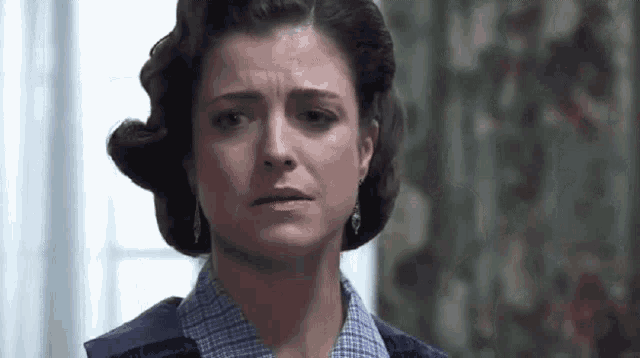a woman with a sad look on her face is standing in front of a window in a room .