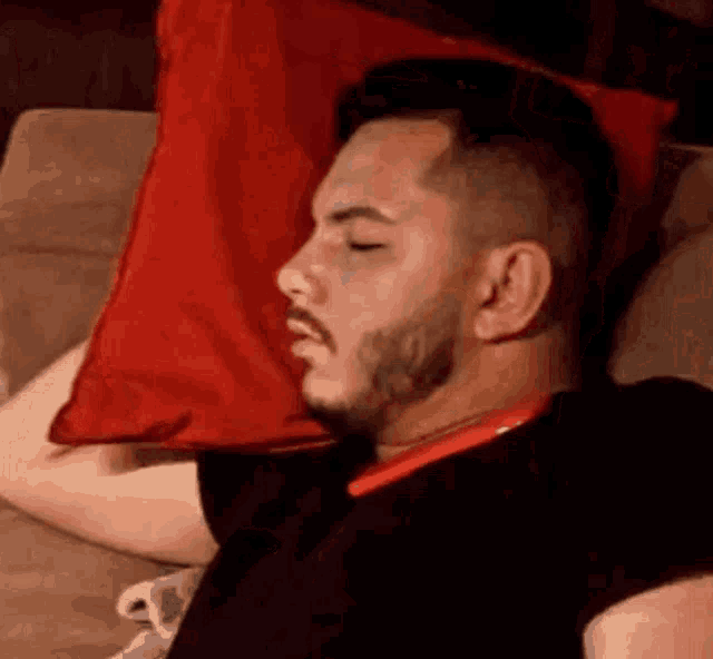 a man with a beard is sleeping on a couch next to a red pillow .