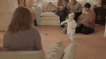 a baby is walking in a living room with a group of people sitting on a couch .