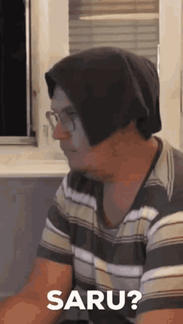 a man wearing a beanie and glasses is sitting in front of a window and says saru .