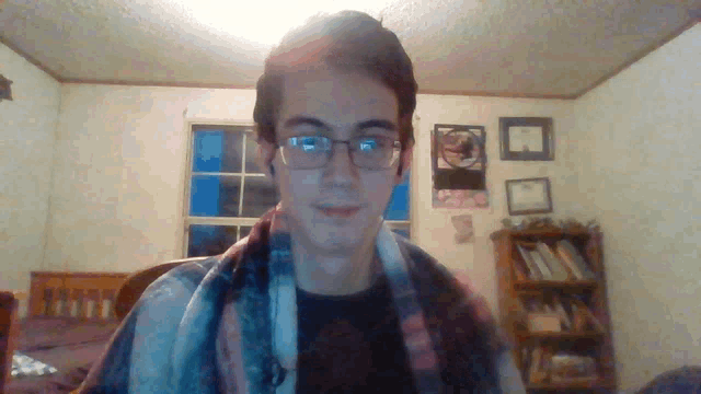 a young man wearing glasses and a plaid shirt looks at the camera in a bedroom