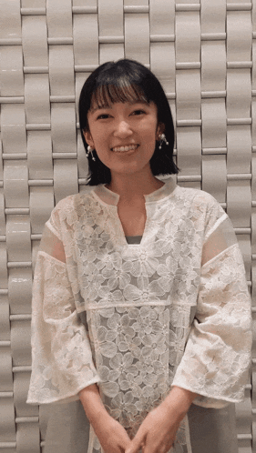a woman wearing a white lace top and earrings smiles in front of a white wall