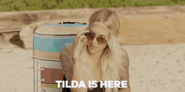 a woman wearing sunglasses is sitting on a beach with the words tilda is here behind her