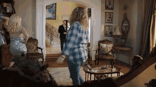 a woman in a plaid shirt is dancing in a living room with a man in a suit behind her .