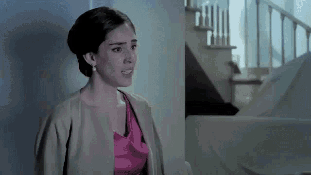 a woman in a pink dress and a tan jacket is standing in front of a staircase in a dark room .