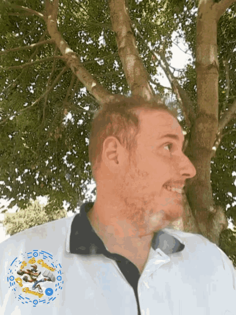 a man stands in front of a tree wearing a white shirt with a blue and yellow emblem on the front
