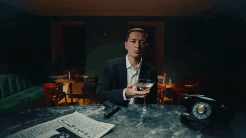 a man sitting at a table with a martini glass and a newspaper that says ' murder ' on it