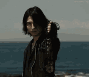 a man with long hair is standing on a beach holding a necklace in his hand .