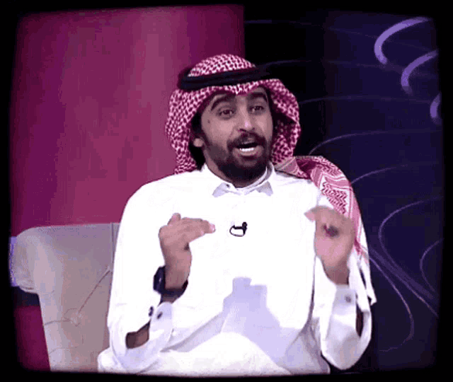 a man with a beard wearing a head scarf and a white shirt has a microphone on his neck
