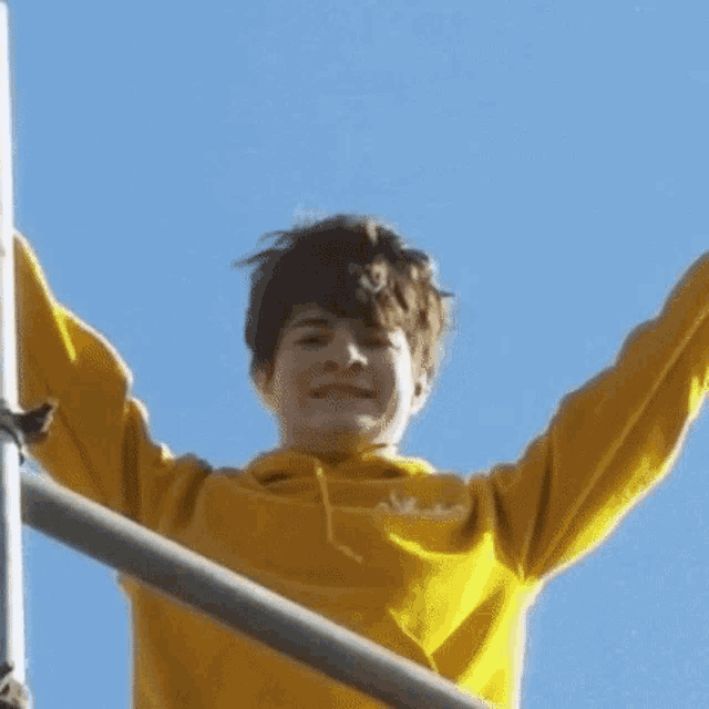a young man wearing a yellow hoodie is standing on a scaffolding with his arms in the air .