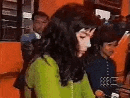 a woman in a green sweater is talking on a telephone while a man stands behind her .