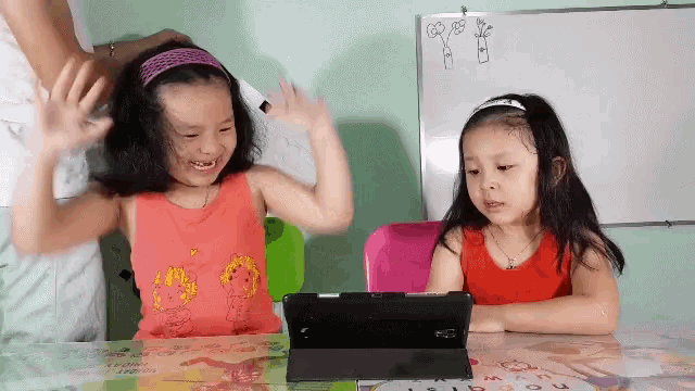 two little girls sitting at a table with a tablet