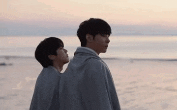 two young men are standing next to each other on a beach looking at the ocean .