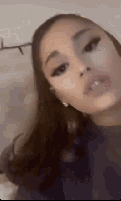 ariana grande is wearing a black shirt and earrings and making a face .