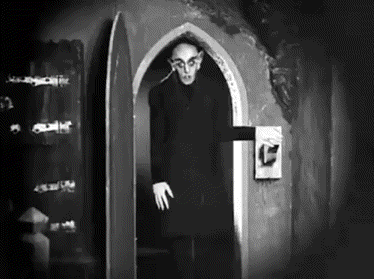 a black and white photo of a vampire standing in a doorway in a dark room .