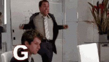 a man in a suit and tie is jumping in the air while another man sits in a chair with the letter g behind him