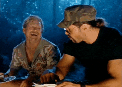 two men are sitting at a table laughing and one is wearing a camouflage hat