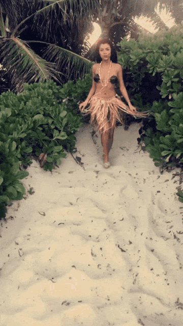 a woman in a bikini and a feathered skirt is walking on a sandy path