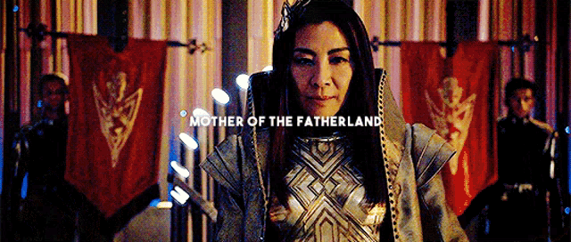 a woman with a crown on her head is standing in front of a group of people and says mother of the fatherland .