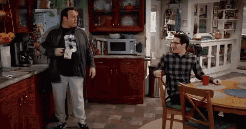 two men are standing in a kitchen talking to each other while a man sits at a table with a cup of coffee .
