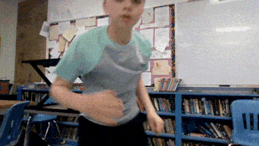 a boy in a classroom with bookshelves and a white board behind him