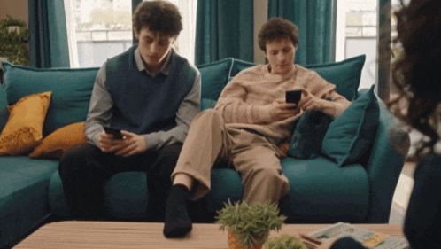 two young men sitting on a blue couch looking at their phones