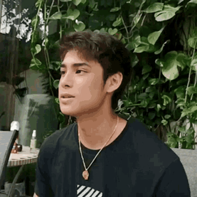a young man wearing a black t-shirt and a gold necklace is sitting in front of a table .