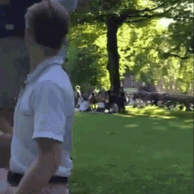 a man in a white shirt stands in a park with people sitting on the grass .