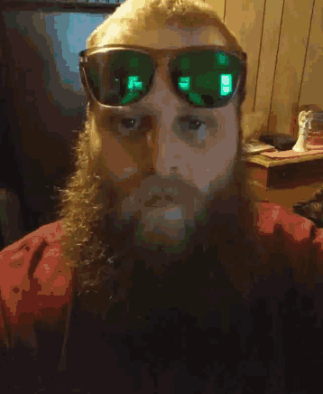 a man with a beard wearing green sunglasses looks at the camera
