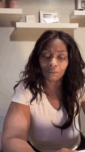 a woman with long hair wearing a white shirt is sitting in front of a shelf