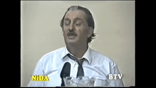 a man in a white shirt and tie is talking into a microphone while holding a glass of water .