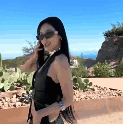 a woman wearing sunglasses and a choker stands in front of a cactus