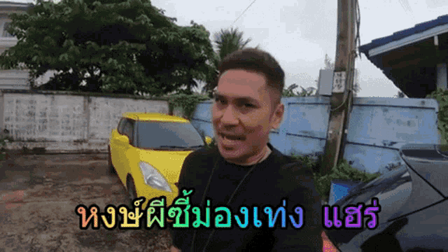 a man in a black shirt stands in front of a yellow car in a parking lot with foreign writing on it