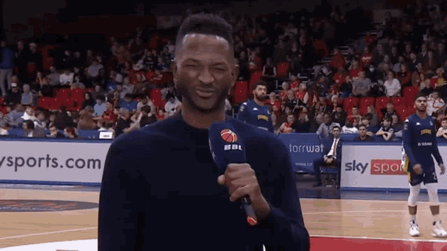 a man holding a microphone on a basketball court with a sky sports sign behind him