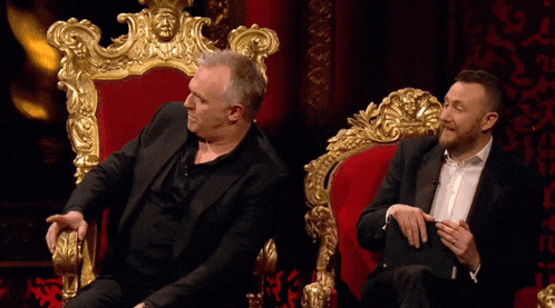 a man in a black suit is sitting on a gold throne