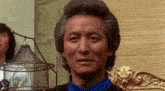 a man with gray hair and a blue shirt is standing in front of a brick wall and looking at the camera .