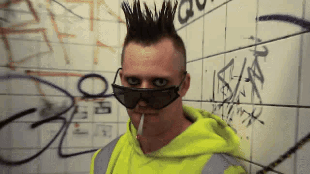 a man wearing sunglasses and a mohawk smoking a cigarette in front of a wall with graffiti on it