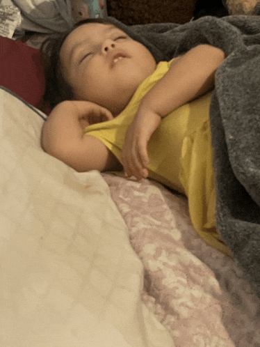 a baby in a yellow tank top is sleeping on a bed under a blanket