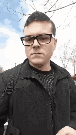 a man wearing glasses and a black jacket is standing in front of trees .
